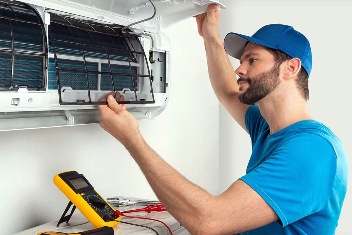 Técnico con gorra azul levantando la tapa de un split de pared para sacar el filtro, con un polímetro amarillo apoyado debajo, en una revisión en Elche