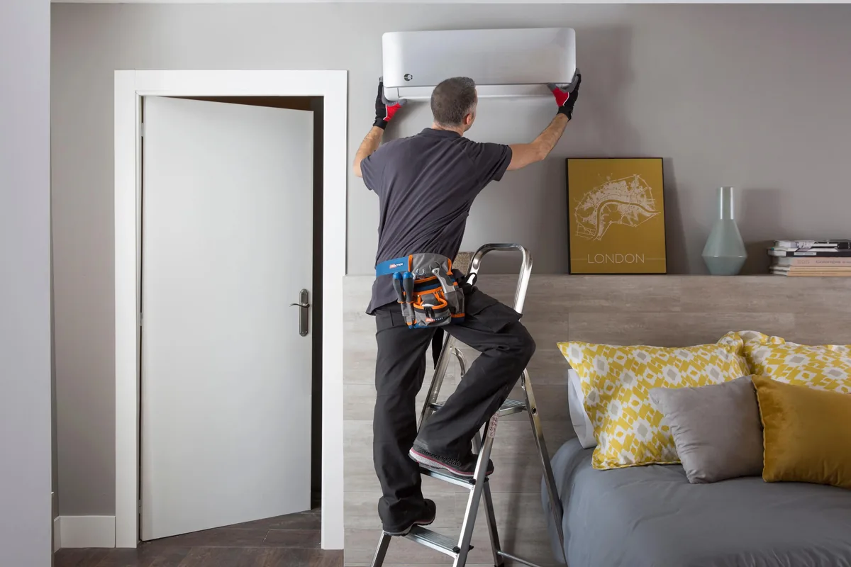 Técnico subido a una escalera sujetando la unidad interior de un split sobre el cabecero de un dormitorio en Málaga
