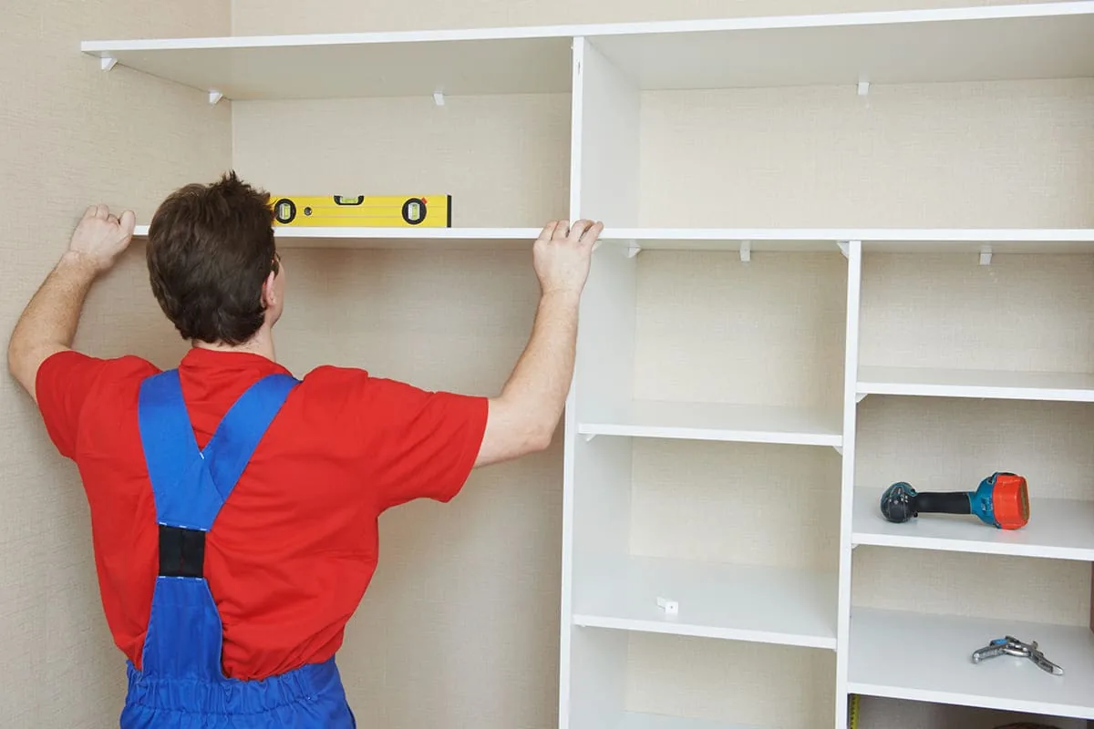 Operario con camiseta roja apoyando un nivel amarillo sobre el estante alto de un armario blanco a medida, con el taladro en una balda, en una vivienda de Zaragoza