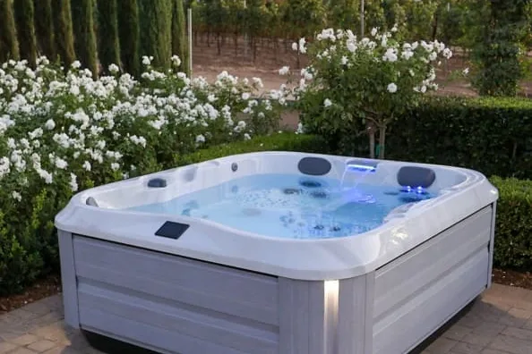 Spa portátil de faldón gris claro con cascada iluminada sobre un patio de adoquín, entre rosales blancos y cipreses, como el de un jardín pareado de Covaresa en Valladolid