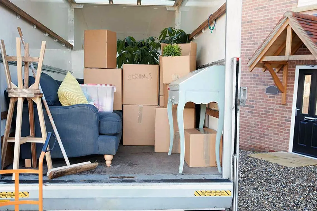 Camión de mudanzas con la puerta trasera abierta y la plataforma bajada, cargado con un sofá azul, cajas, plantas y una mesa auxiliar, frente a una vivienda en Alicante