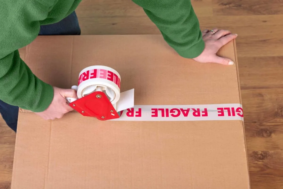Manos con jersey verde precintan una caja de cartón con cinta roja y blanca de la palabra fragile sobre un suelo de madera, preparando una mudanza en Elche