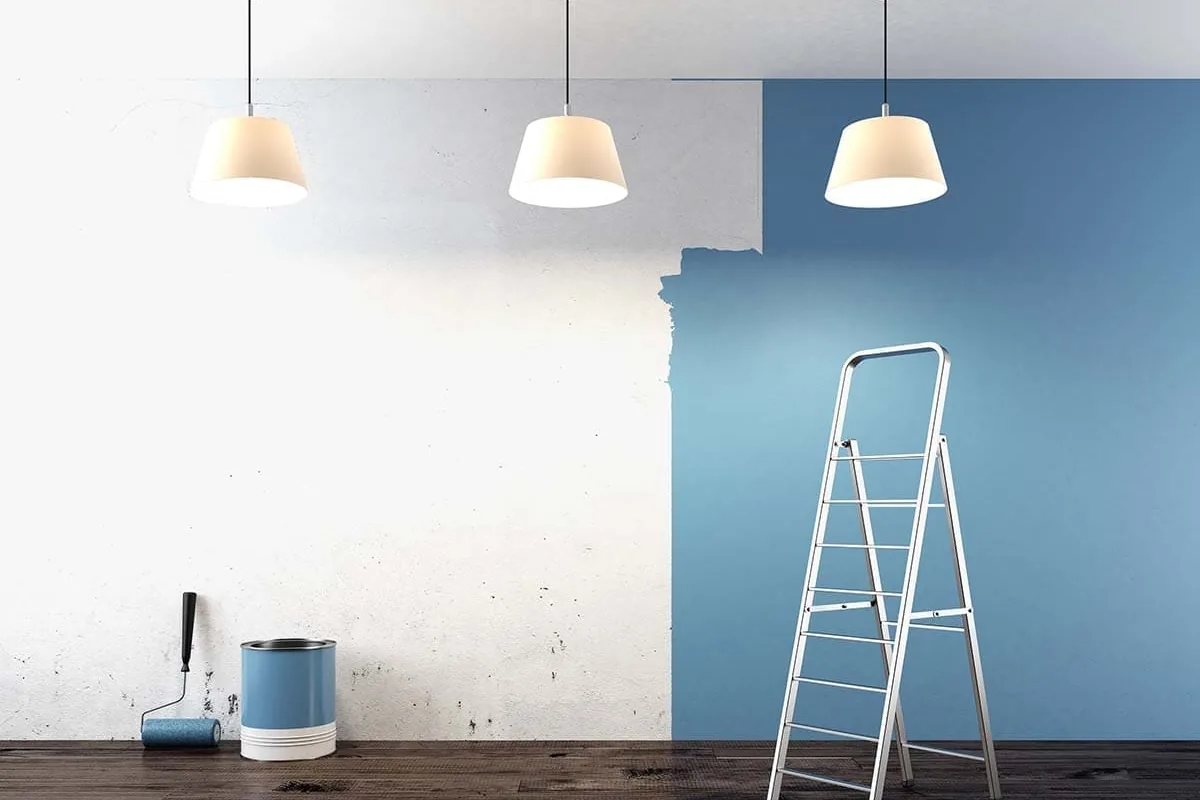 Pared de un salón en Sevilla a medio pintar, con el enlucido viejo manchado junto a un paño azul, rodillo, bote y escalera de tijera