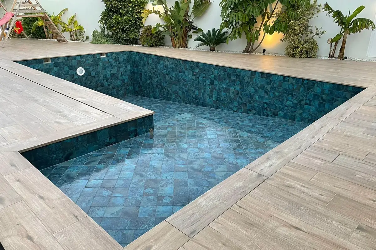 Piscina de obra recién revestida con baldosa cerámica verde azulada y aún sin agua, rodeada de tarima porcelánica efecto madera en el jardín de un chalé de Mallorca