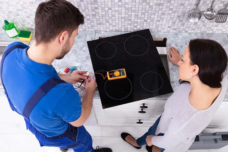 Vista cenital de un técnico con mono azul y polímetro comprobando los cables de una placa de inducción negra apoyada sobre una encimera de piedra, con una clienta a su lado, en una cocina de Mallorca