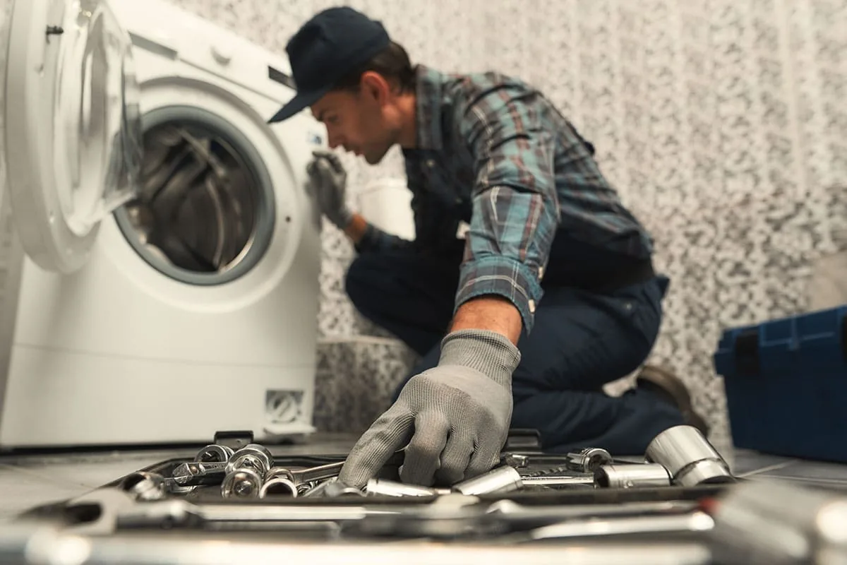 Técnico con gorra y camisa de cuadros arrodillado ante una lavadora con la puerta abierta, con una bandeja de vasos y llaves de tubo en primer plano, en un baño de un piso de Vigo