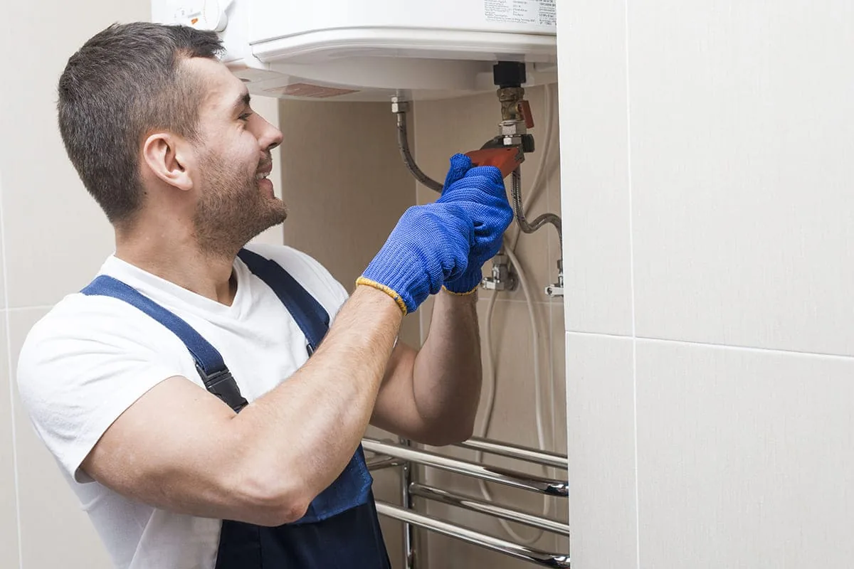 Profesional con guantes azules apretando con una llave grifa las conexiones bajo un termo eléctrico blanco, junto a un toallero, en un baño alicatado de una vivienda de Elche