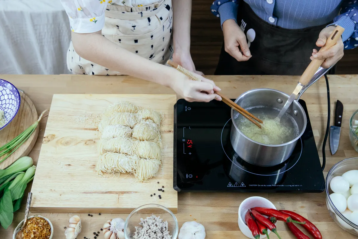 Amas de casa con delantal hirviendo fideos en una placa de sobremesa en una cocina moderna