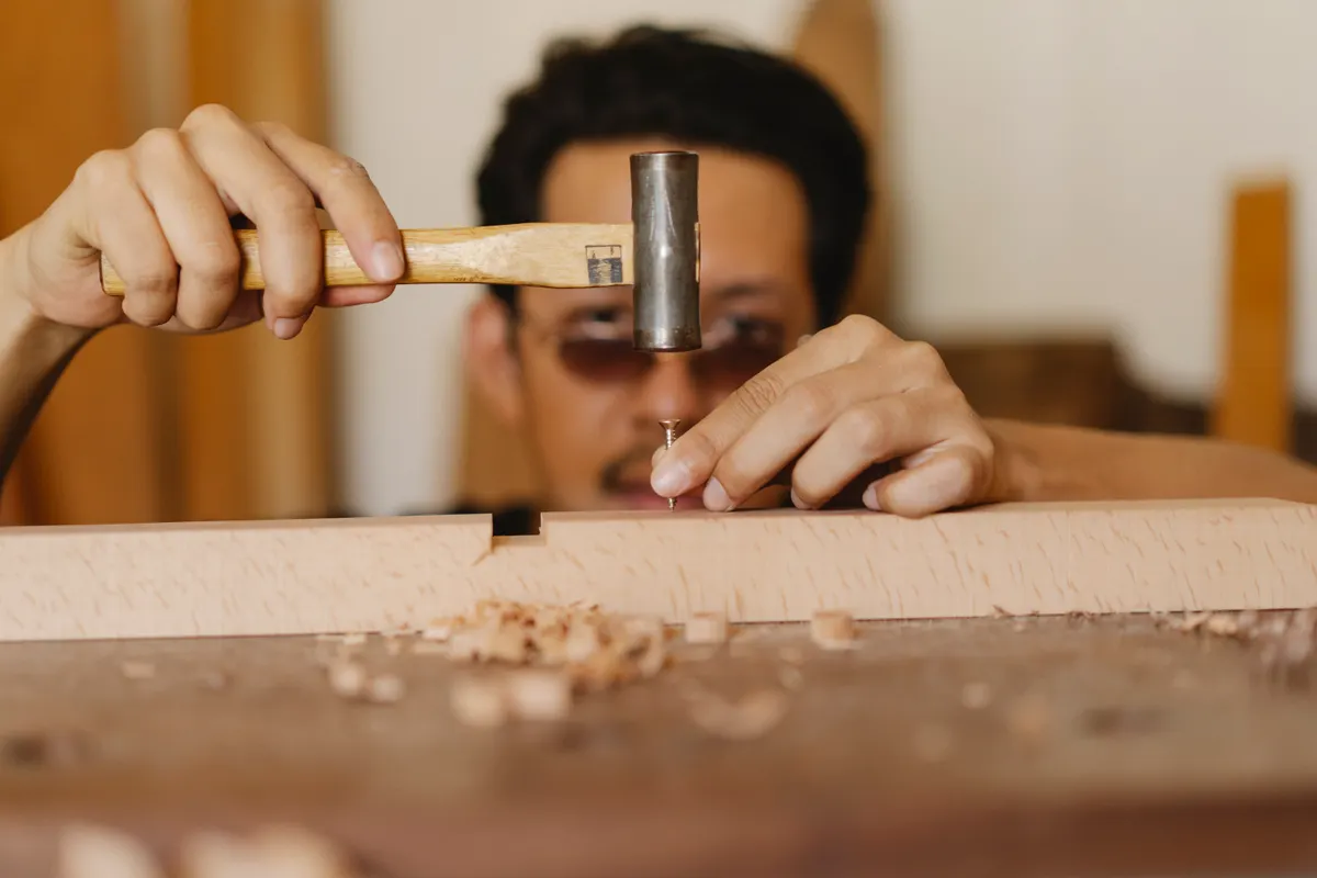 Artesano joven con gafas clavando un clavo en un tablón en un taller de carpintería
