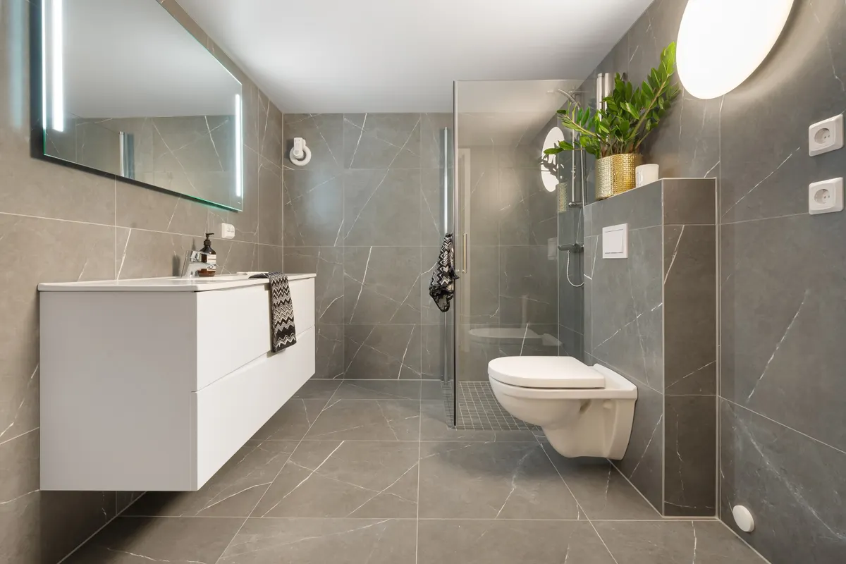 Baño de azulejo gris con ducha, inodoro y espejo iluminado