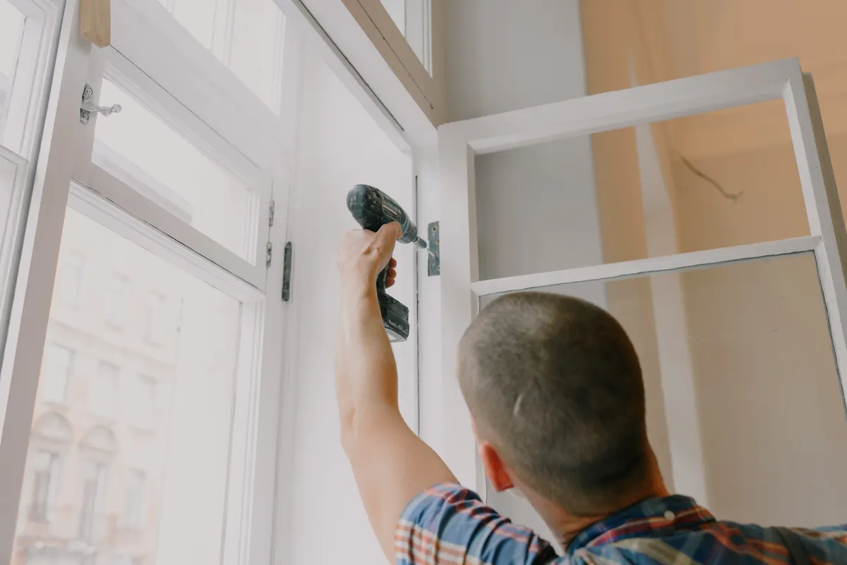 Carpintero fijando con un atornillador inalámbrico la bisagra de una ventana de madera blanca en un piso