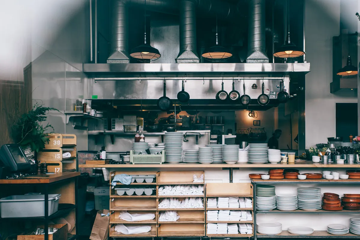 Cocina de restaurante con campanas de acero, sartenes colgadas y pilas de platos