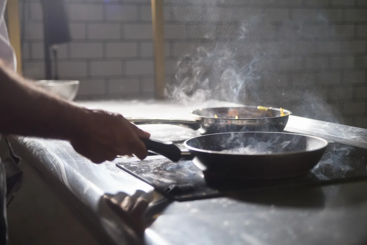 Cocinero usando varias sartenes sobre una placa eléctrica en una cocina llena de vapor