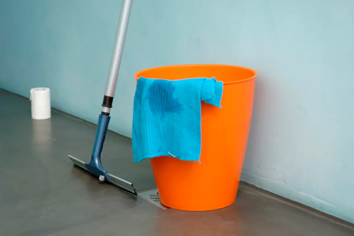 Cubo naranja con una bayeta azul en el borde y una rasqueta de goma para el suelo apoyada a su lado