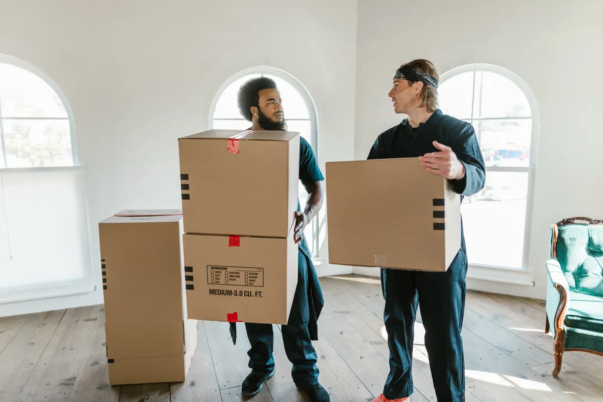 Dos operarios de mudanzas con mono de trabajo llevan cajas de cartón por una vivienda vacía y luminosa