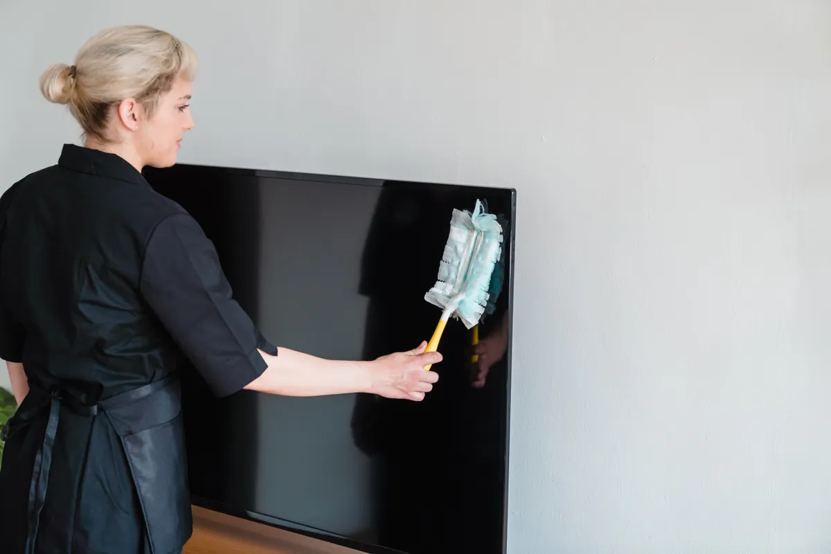 Limpiadora de uniforme negro quitando el polvo de una pantalla de televisión apagada con un plumero