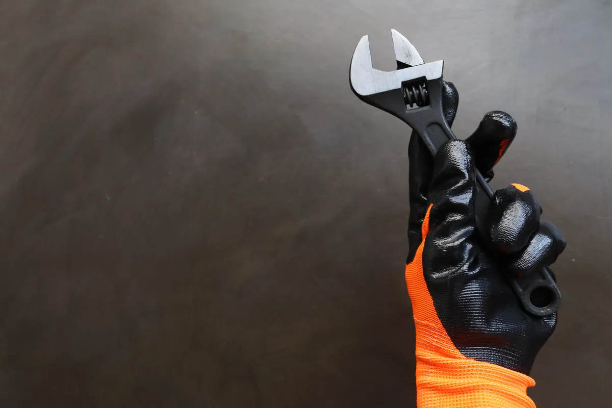 Mano con guante de trabajo naranja y negro sujetando una llave inglesa sobre un fondo oscuro