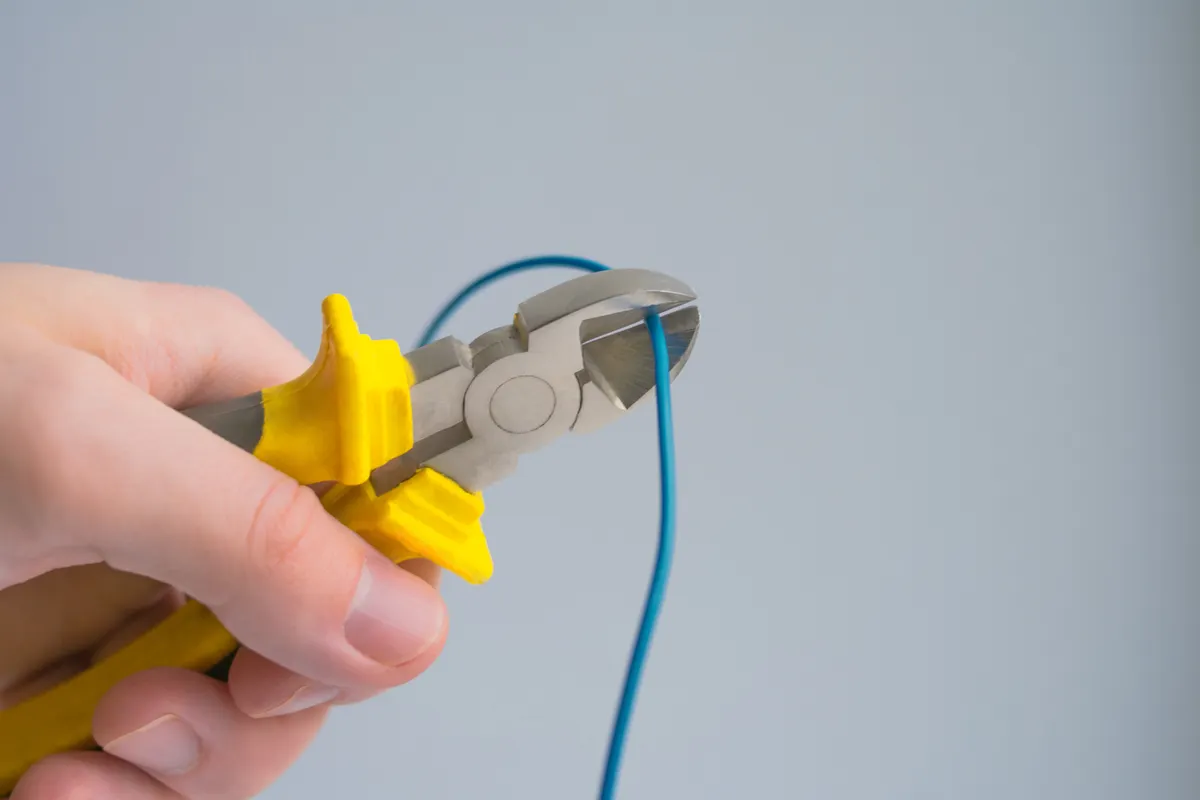 Mano cortando un cable azul con unos alicates sobre un fondo gris
