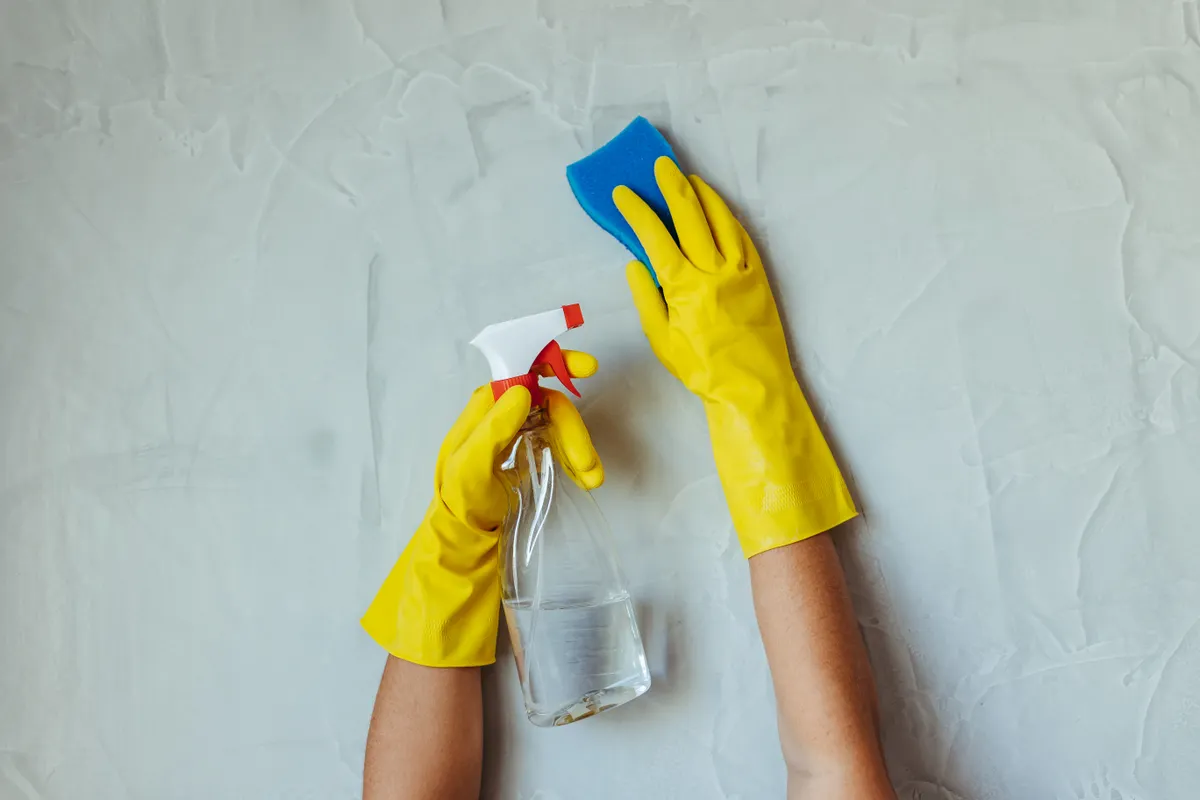 Manos con guantes de goma amarillos sujetando un pulverizador y una esponja azul sobre una superficie gris