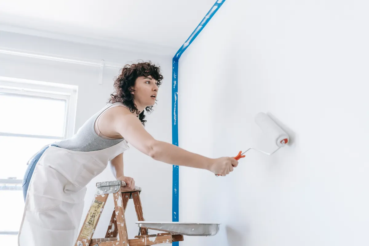 Mujer subida a una escalera pintando una pared de blanco en una reforma casera