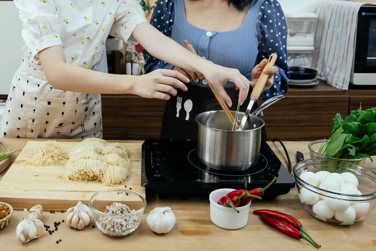 Mujeres con delantal cociendo fideos caseros en un cazo sobre una placa de sobremesa rodeada de ingredientes frescos