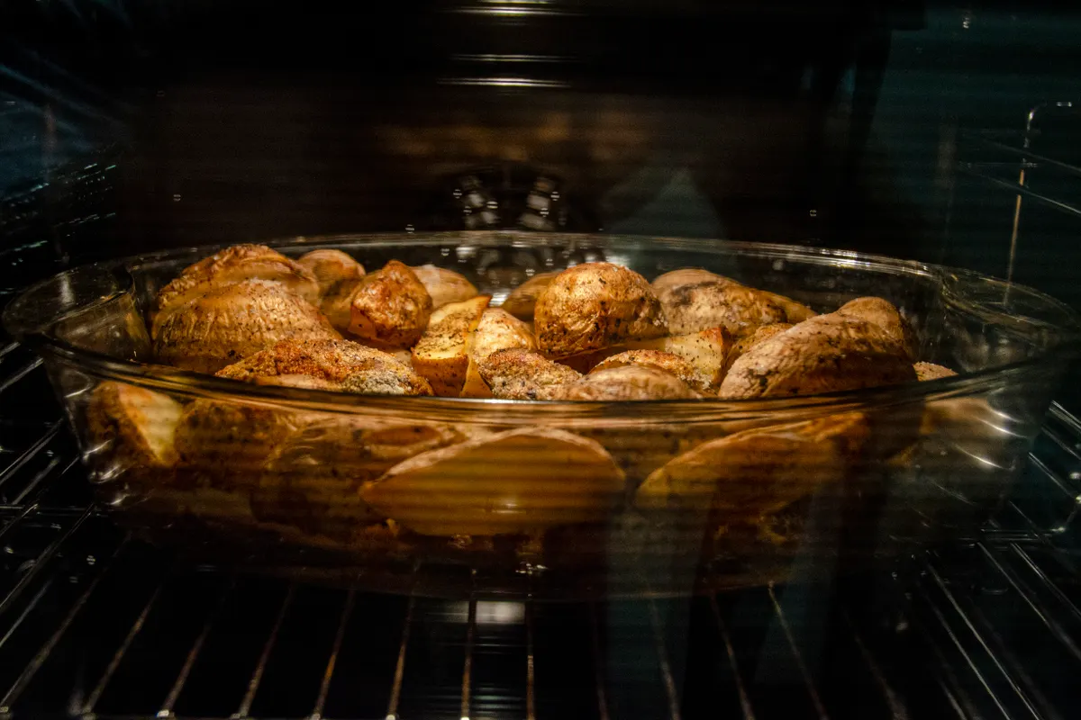 Patatas con piel y especias asándose en una fuente, vistas a través del cristal del horno