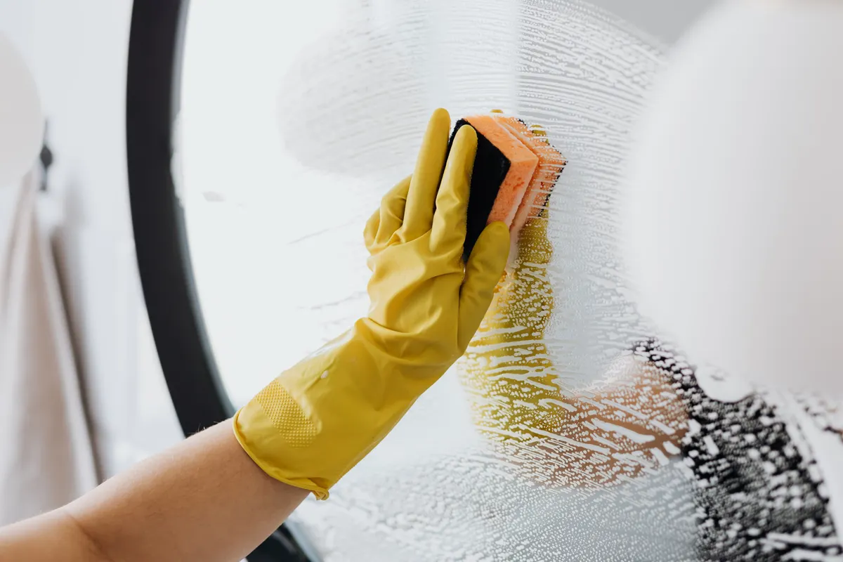 Persona con guantes de goma amarillos limpiando el espejo del baño con una esponja