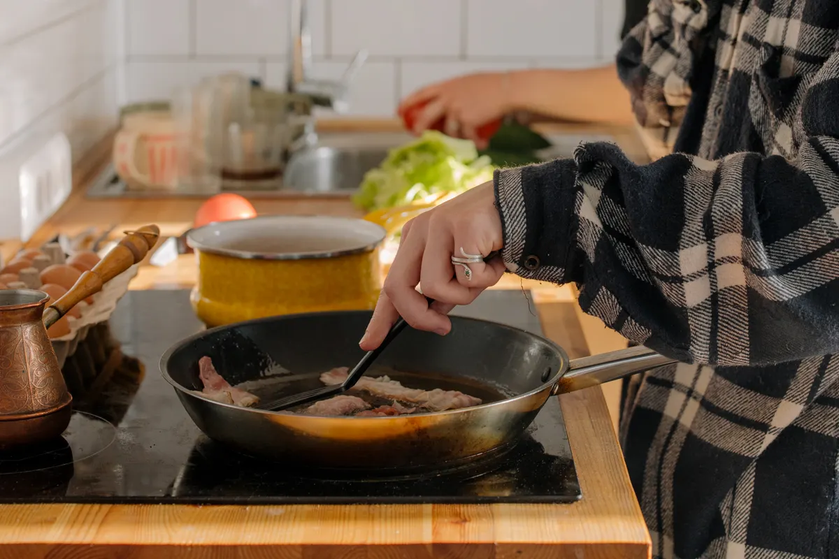Persona friendo beicon en una sartén en una cocina acogedora con ingredientes frescos al lado